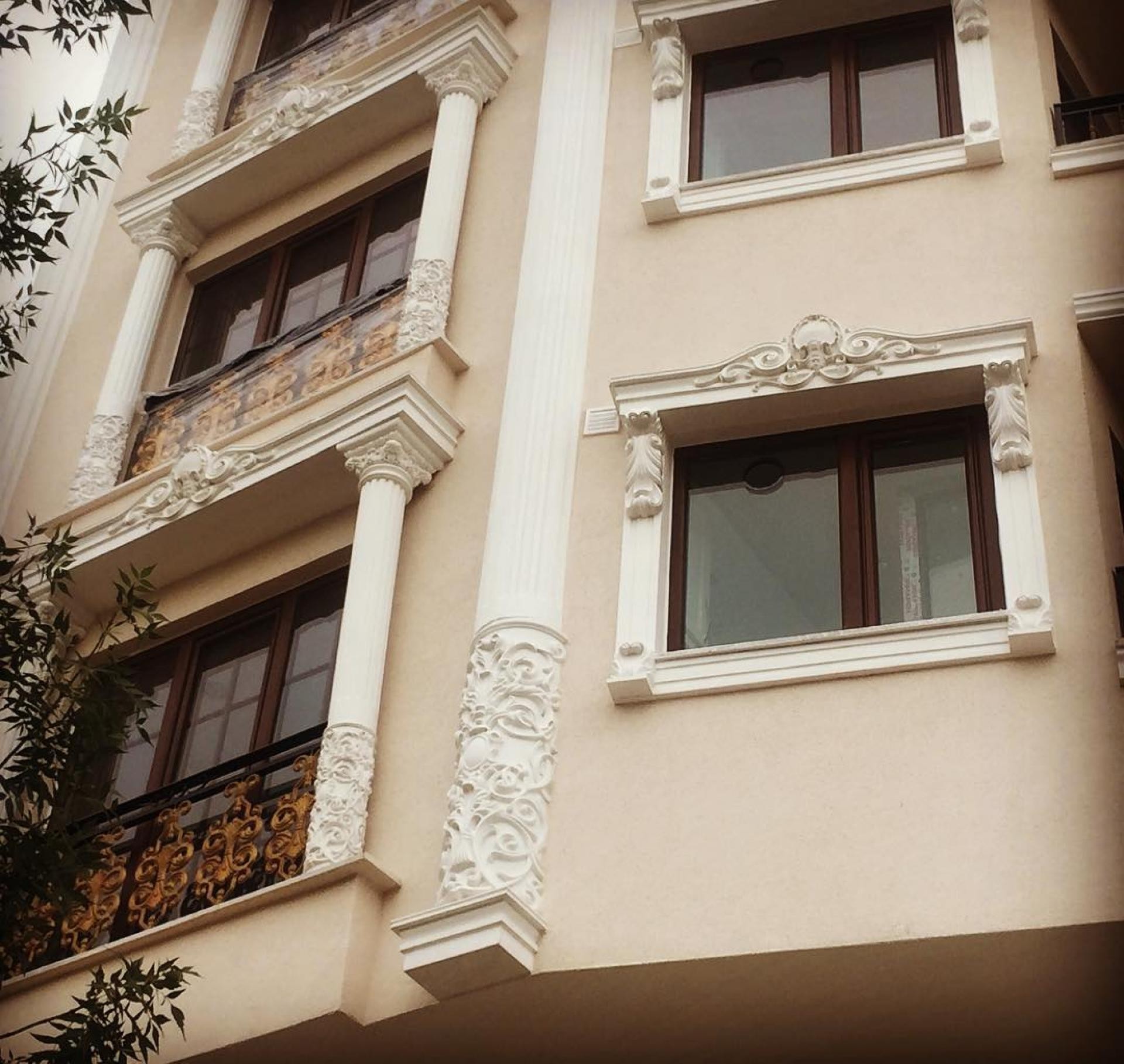 Krem rengi bir binanın dış cephesinde yer alan beyaz renkli dekoratif sütunlar, pencere söveleri ve altın sarısı detaylı balkon korkulukları.
