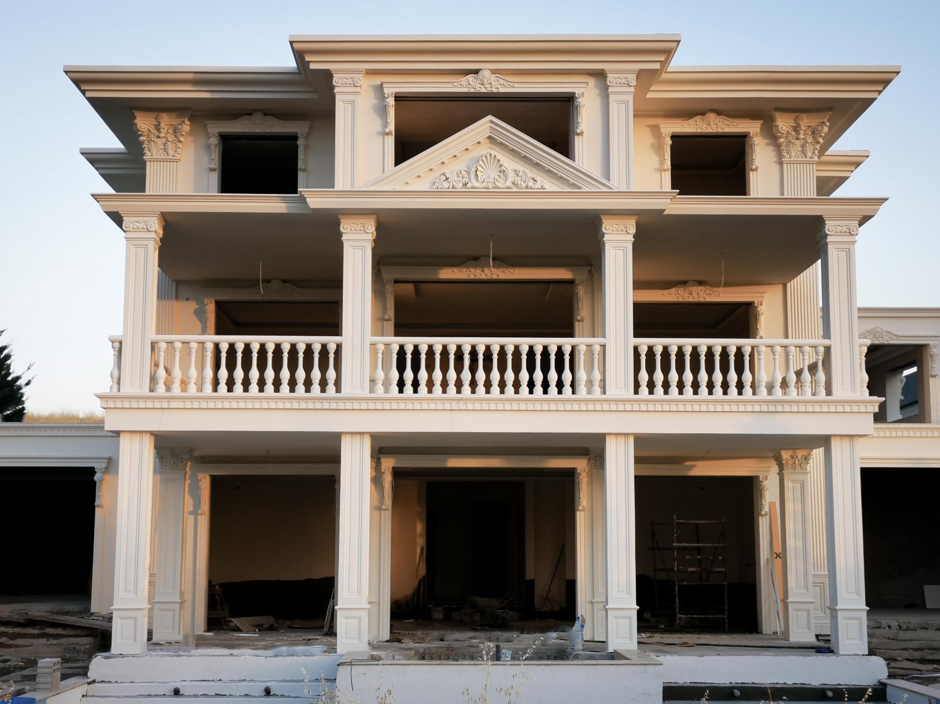 Yapım aşamasında olan, ön cephesinde beyaz renkli poliüretan kare sütunlar, geniş balkon korkulukları, pencere söveleri ve büyük bir alınlık bulunan üç katlı villa.