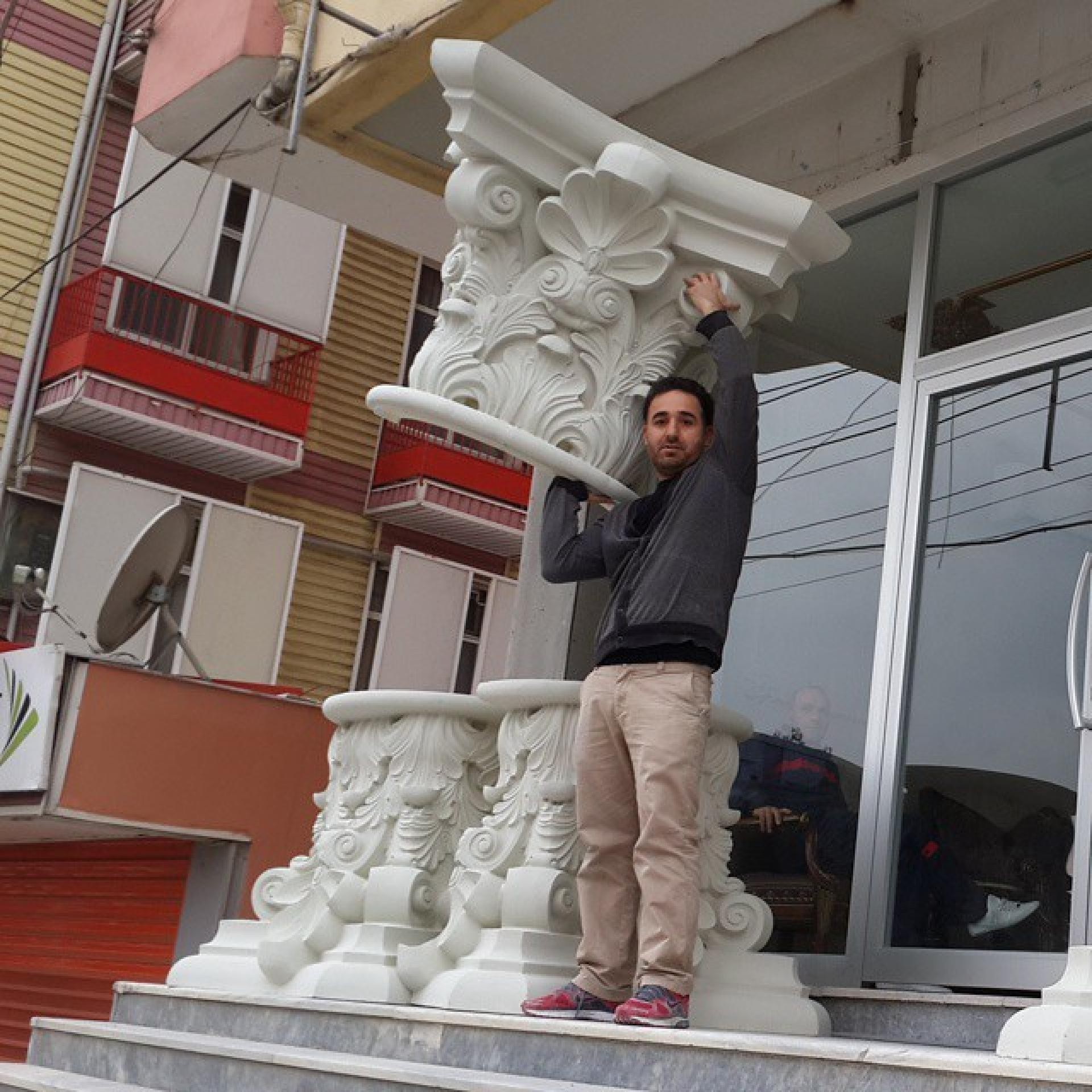 Bir binanın önündeki merdivenlerde, devasa boyuttaki işlemeli beyaz poliüretan sütun başlığını hafifliği sayesinde tek eliyle omzunda tutan bir adam.