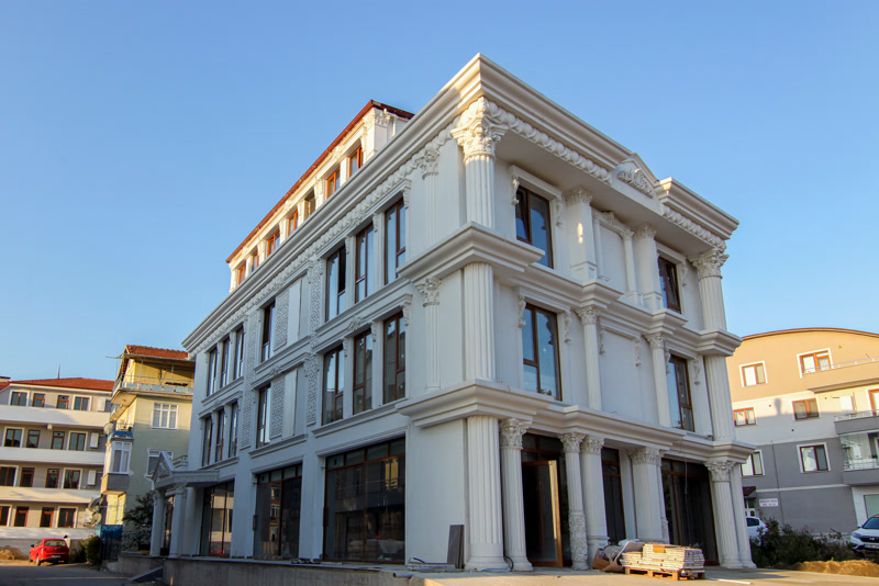 Tamamı beyaz renge boyanmış çok katlı bir binanın dış cephesinde, katlar boyunca uzanan yivli poliüretan pilaster sütunlar, pencere söveleri ve dekoratif oymalı paneller.