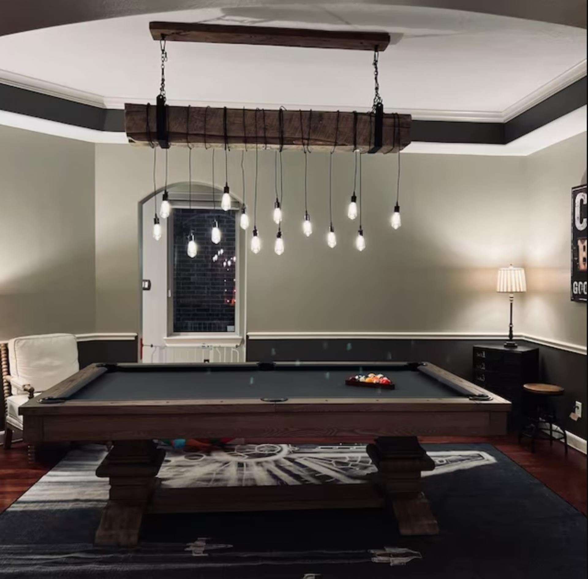 Bilardo masasının üzerinde zincirlerle asılı duran, çok sayıda Edison ampulün bağlandığı ahşap görünümlü dekoratif kütük avize ve gri duvarlı oyun odası.