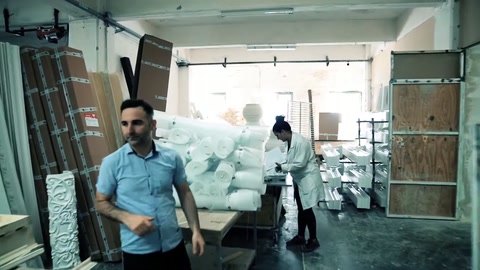 Mavi tişörtlü çalışan, dolu üretim bölümünde kameraya doğru yürüyor.